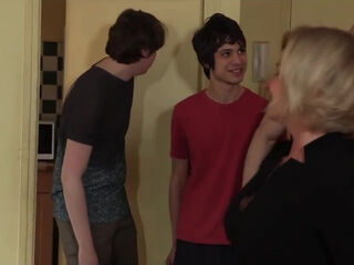 A sophisticated, fair-haired woman engages in group intimacy while an elderly gentleman observes the scene.